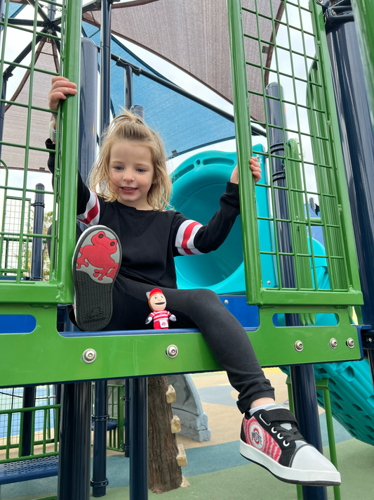 Child sitting wearing bobbi toads shoes with ohio state design and mascot brutus on a playground structure with a slide in the background
