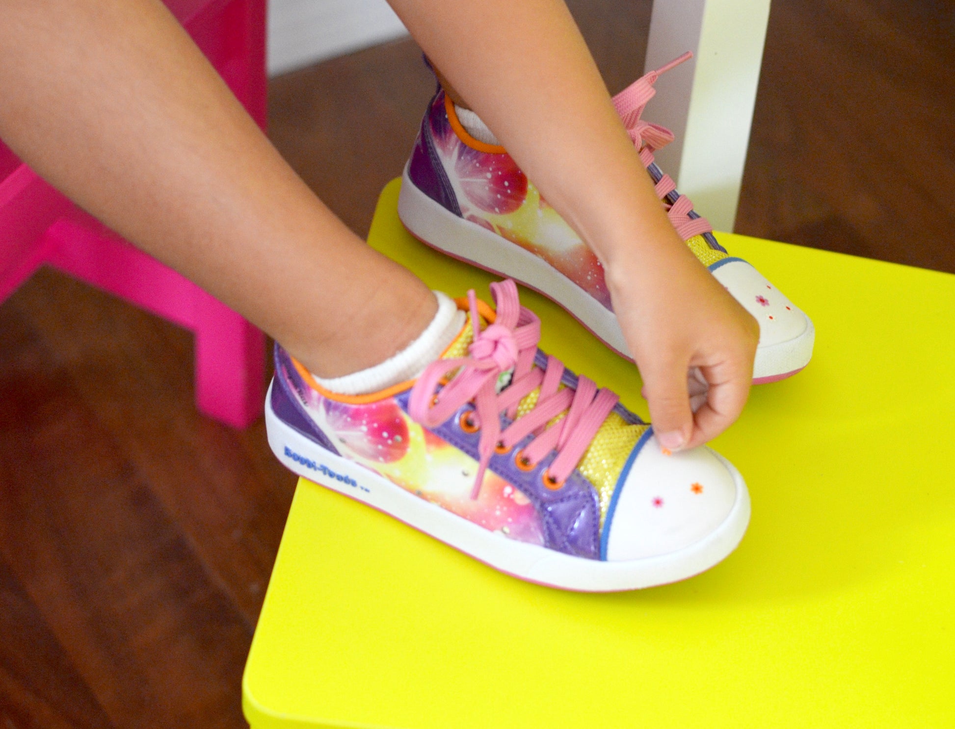 Child wearing colorful sneakers with pink laces on a yellow stool.
