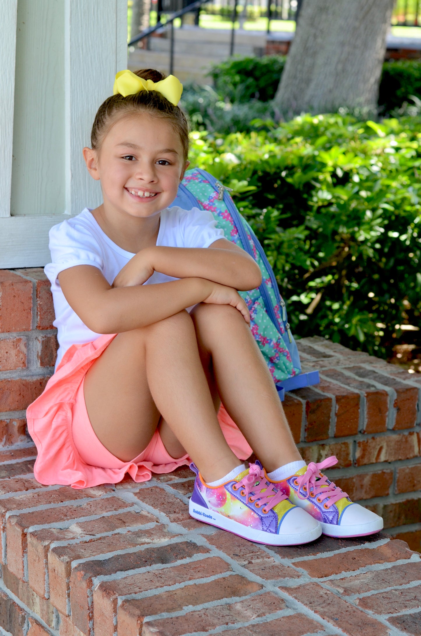 Young girl sitting on a brick step with a colorful backpack, wearing a pink skirt and multicolored sneakers.