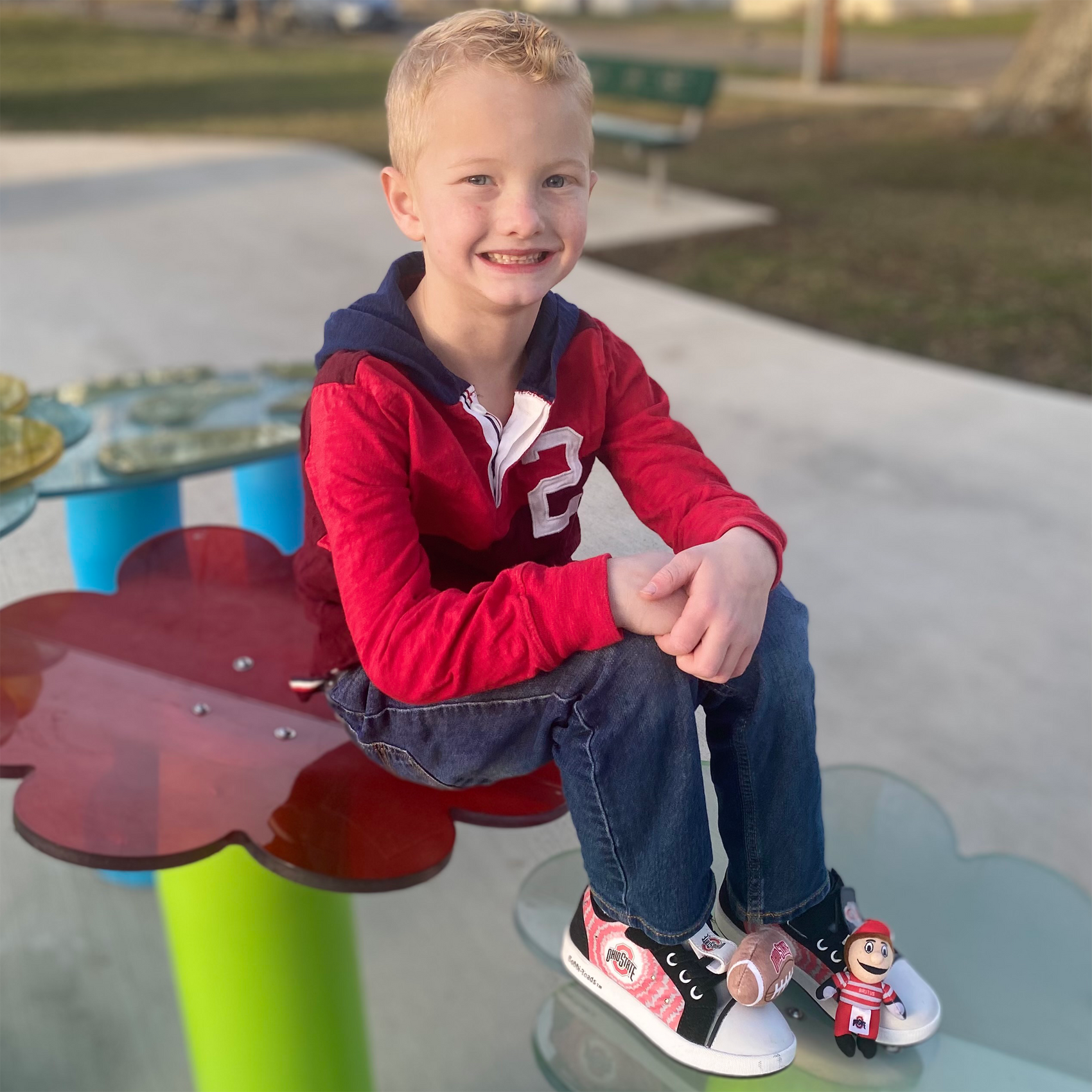 Young boy sitting wearing bobbi-toads ohio state university shoes with mascot brutus bobbi-dobbiez attachable plush charm on a colorful playground structure with a park in the background