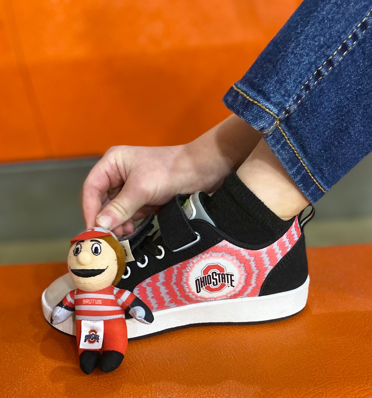 Person wearing a Ohio State-themed sneaker with a brutus bobbi-dobbiez plush charm attached, on an orange surface.