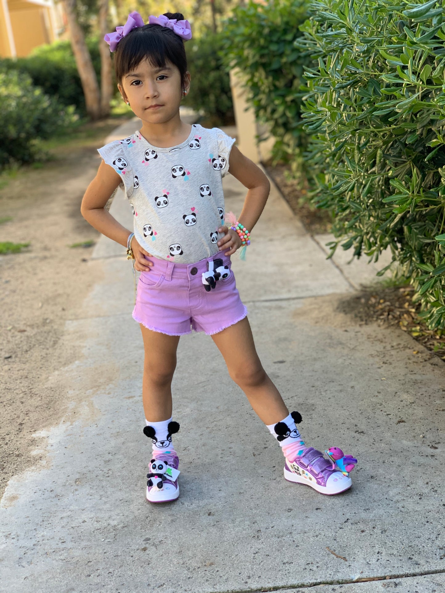 Young girl in a white shirt with black and purple patterns, pink shorts, and matching shoes standing on a sidewalk.